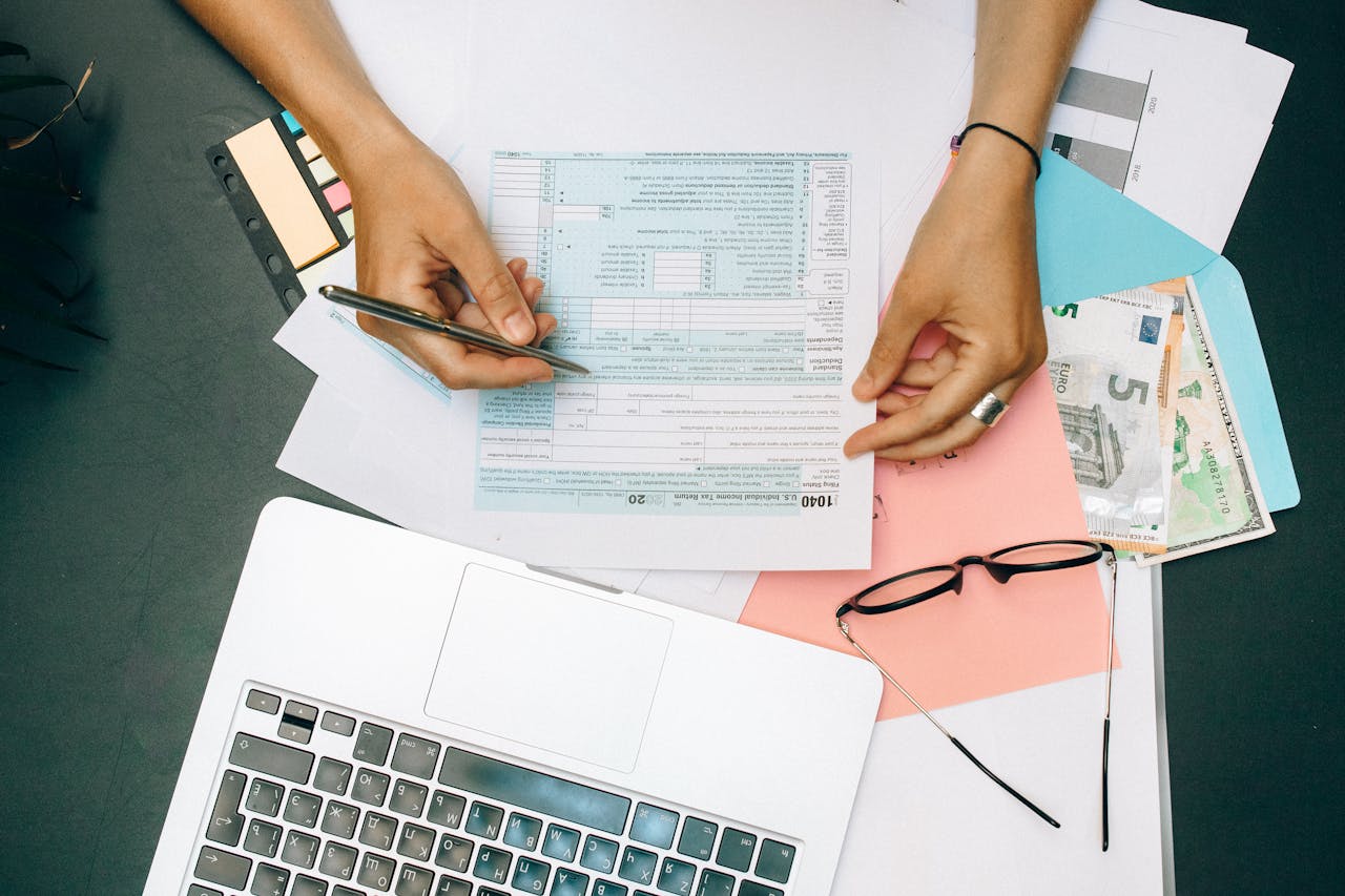 home-hero Hands writing on tax documents with laptop, glasses, and currency on desk.